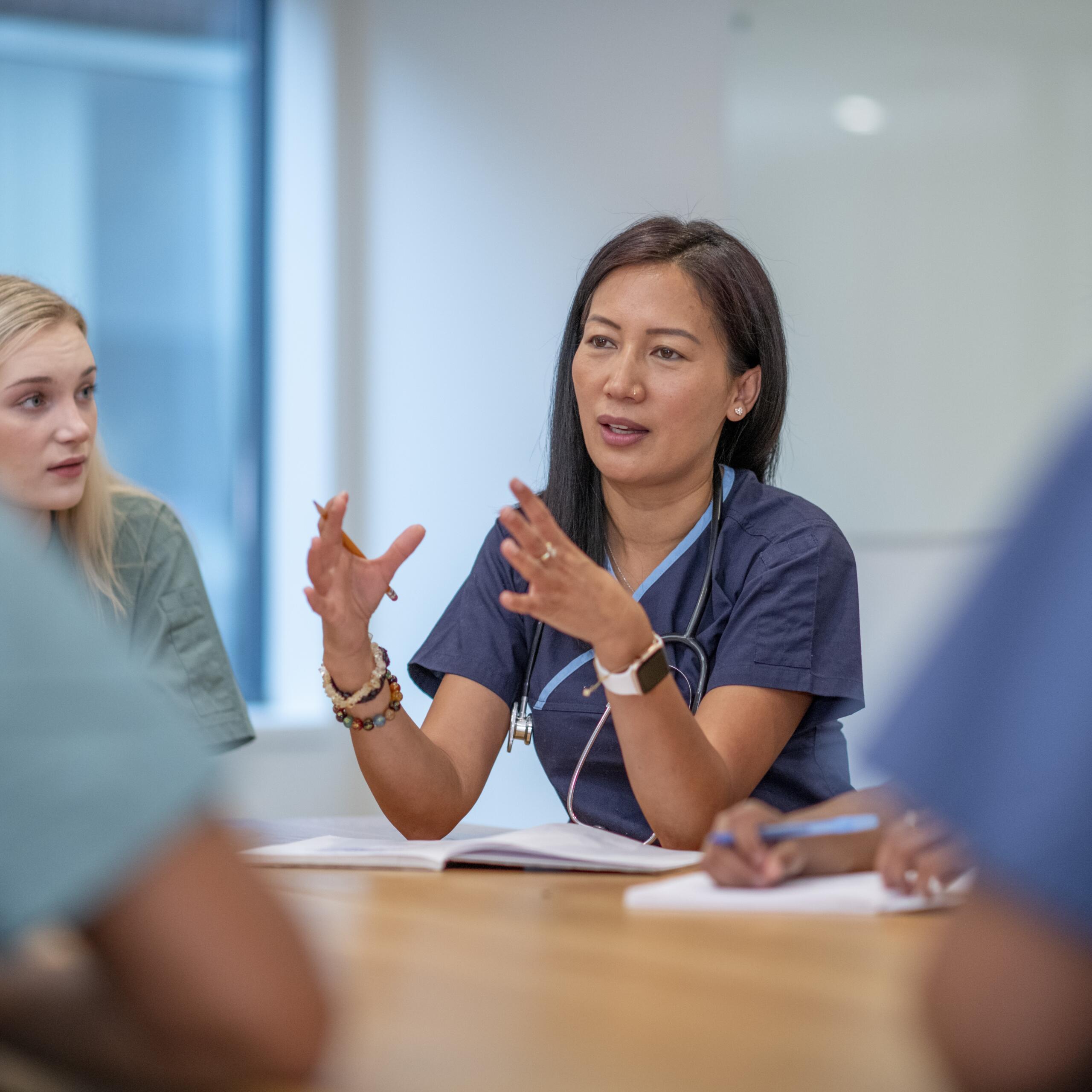 A small group of Nurses sit at a boardroom table as they meet to discuss patient cases. They are each dressed professionally in scrubs and have stethoscopes around their necks as they discuss plans of care for each case.