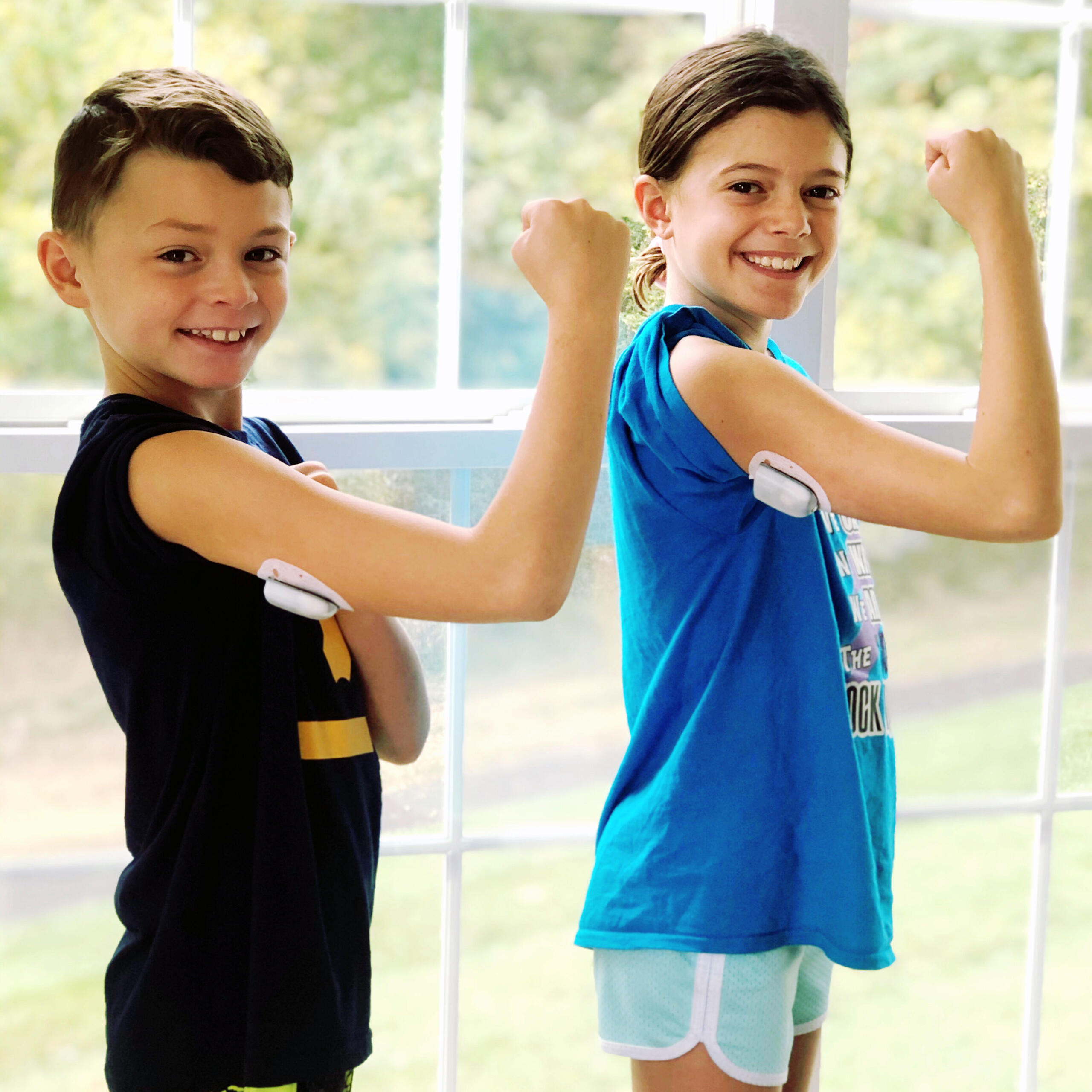 A young boy in a black shirt and a young girl in a blue shirt holding up their arms to show their CGMs