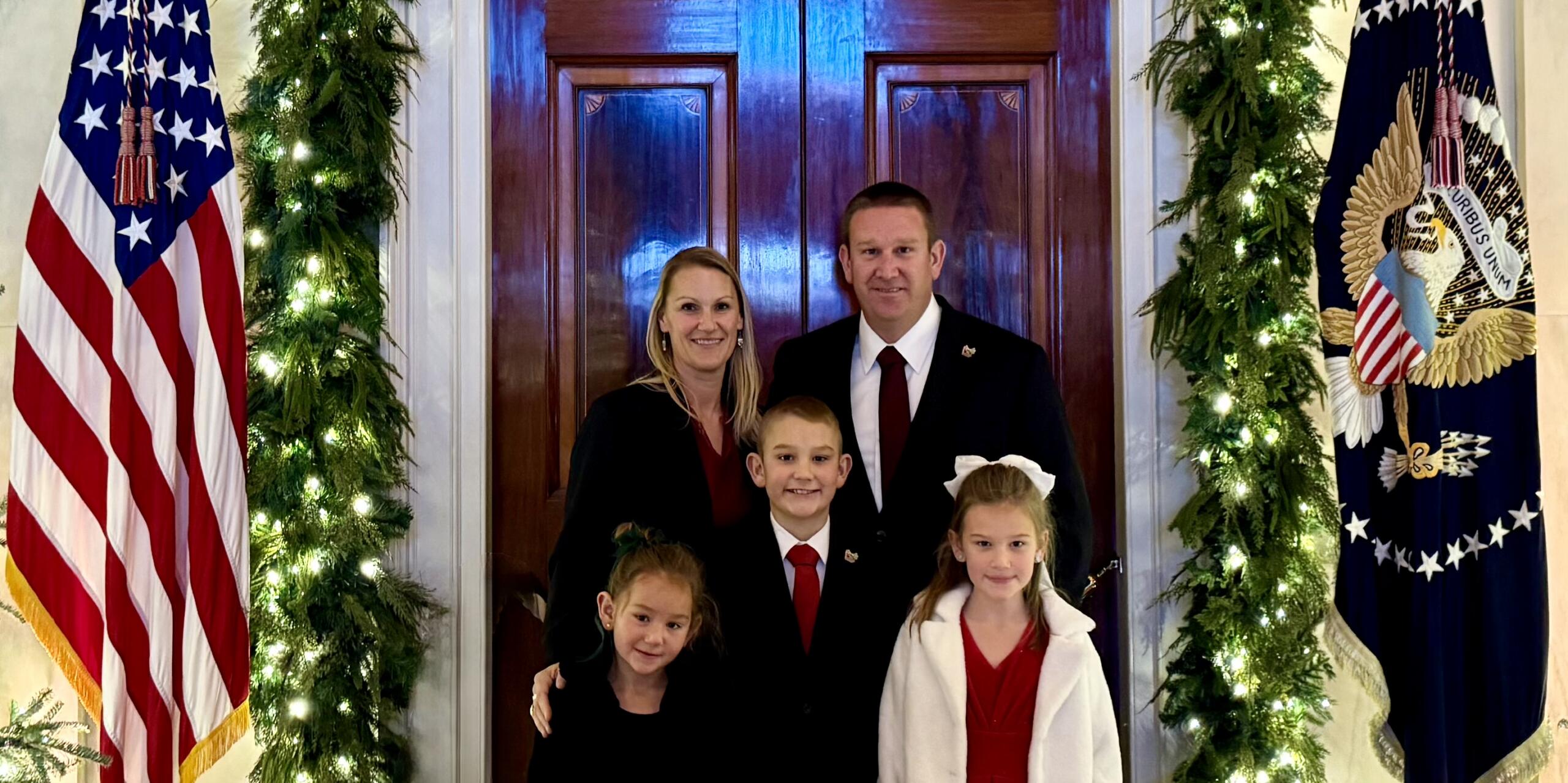 A family of a mother, father, their three children standing next to the American flag and presidential flag