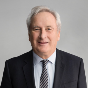 A smiling man in a suit with a light blue shirt and striped tie, set against a neutral gray background.
