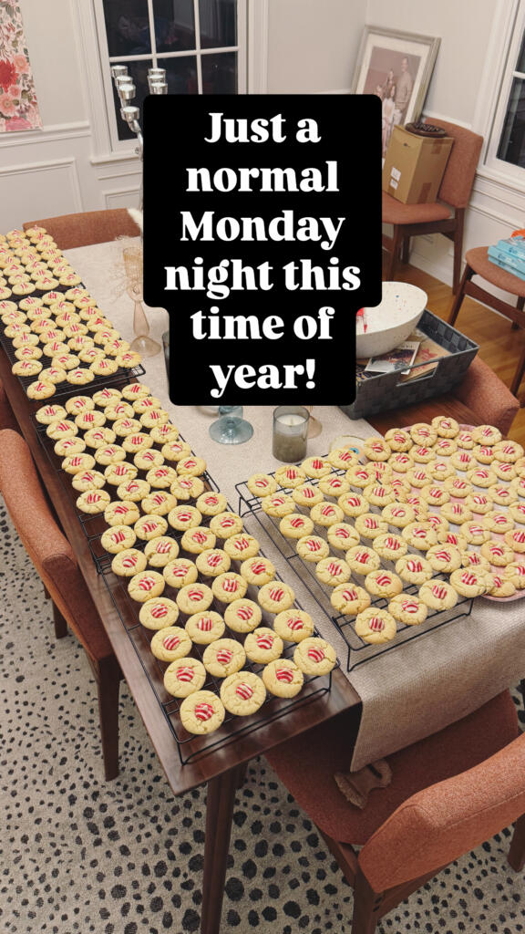 Tons of white cookies with reddish-pink centers lie on cooling racks on a large dining room table. Text above them reads "Just a normal Monday night this time of year!"