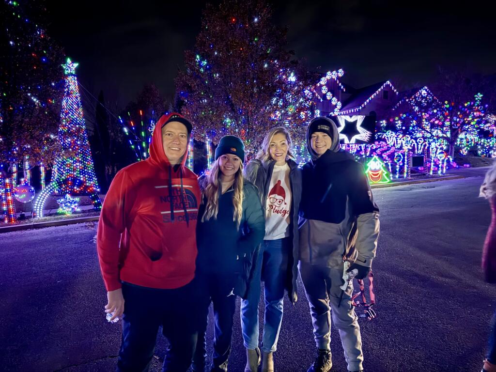 Four white people in winter clothes smile while standing next to each other in front of an elaborate Christmas light display.
