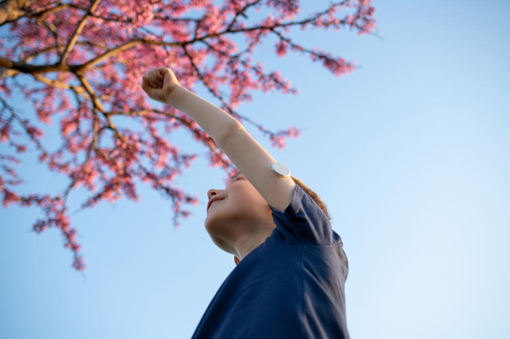 Young boy with type 1 diabetes wears a continuous glucose monitor on his arm and reaches up towards a tree