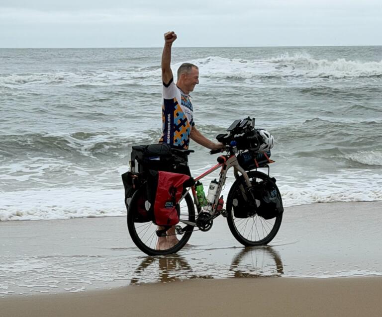 A white man standing next to a bicycle laden with saddlebags on a beach raises his fist in celebration as waves crash behind him.