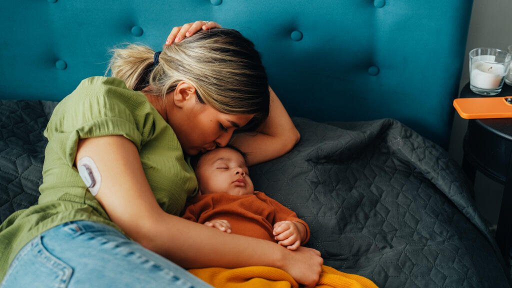 Young mother with type 1 diabetes wearing a continuous glucose monitor (CGM) on her arm sleeping with her baby boy in bed