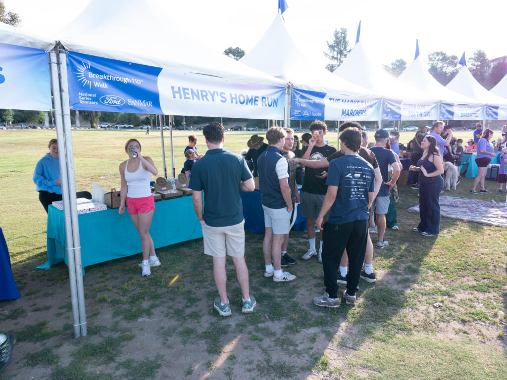 A group of people stand around outside a tent reading "Breakthrough T1D Walk: Henry's Home Run." The tent is next to numerous other tents representing different Walk teams.