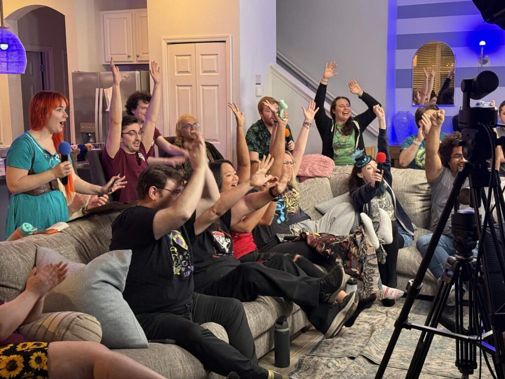 A large group of people sitting on a couch throw their hands in the air in celebration while playing and livestreaming a video game.