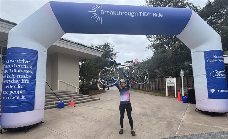 A white woman holds a bicycle over her head in celebration under a large inflatable finish line structure reading "Breakthrough T1D Ride."