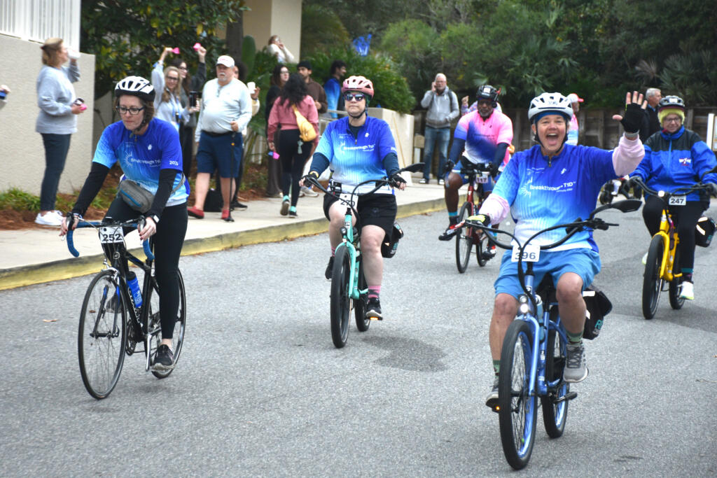 Six people on bikes wearing Breakthrough T1D attire ride down a city street as people on the sidewalk cheer them on.