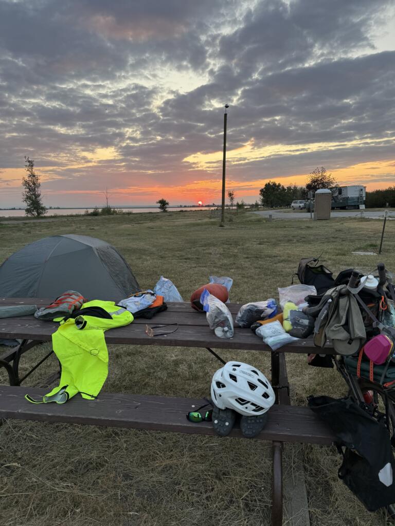 Bike and camping supplies spread out on a picnic table with a sunset in the background.