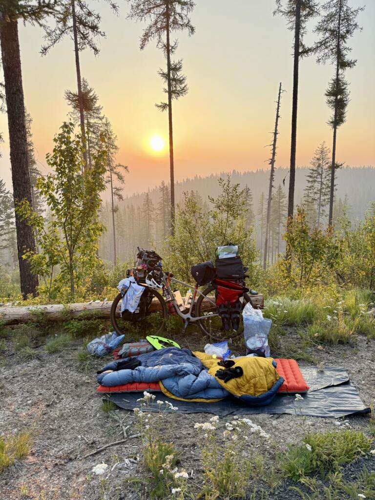 A sleeping bag lies on the ground next to a bike with a sunset and trees in the background.