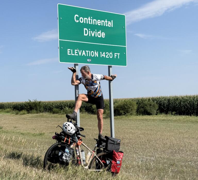 A white man balances on top of a bike covered in bags and panners next to a sign reading "Continental Divide: Elevation 1420 FT."