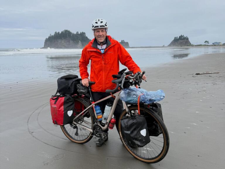 A white man in a red raincoat and white bike helmet smiles while standing next to a bike covered in saddle bags on a beach.