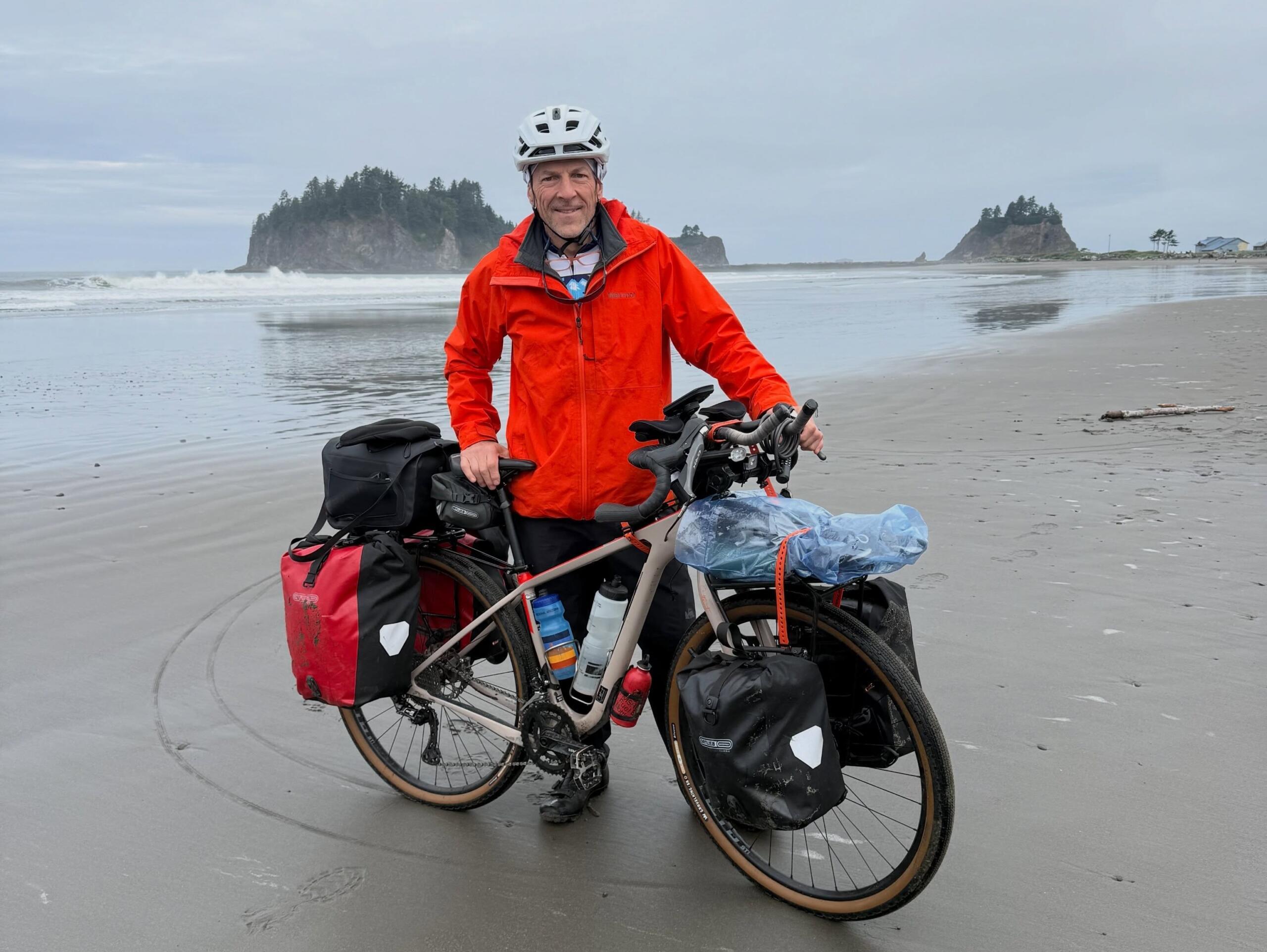 A white man in a red raincoat and white bike helmet smiles while standing next to a bike covered in saddle bags on a beach.