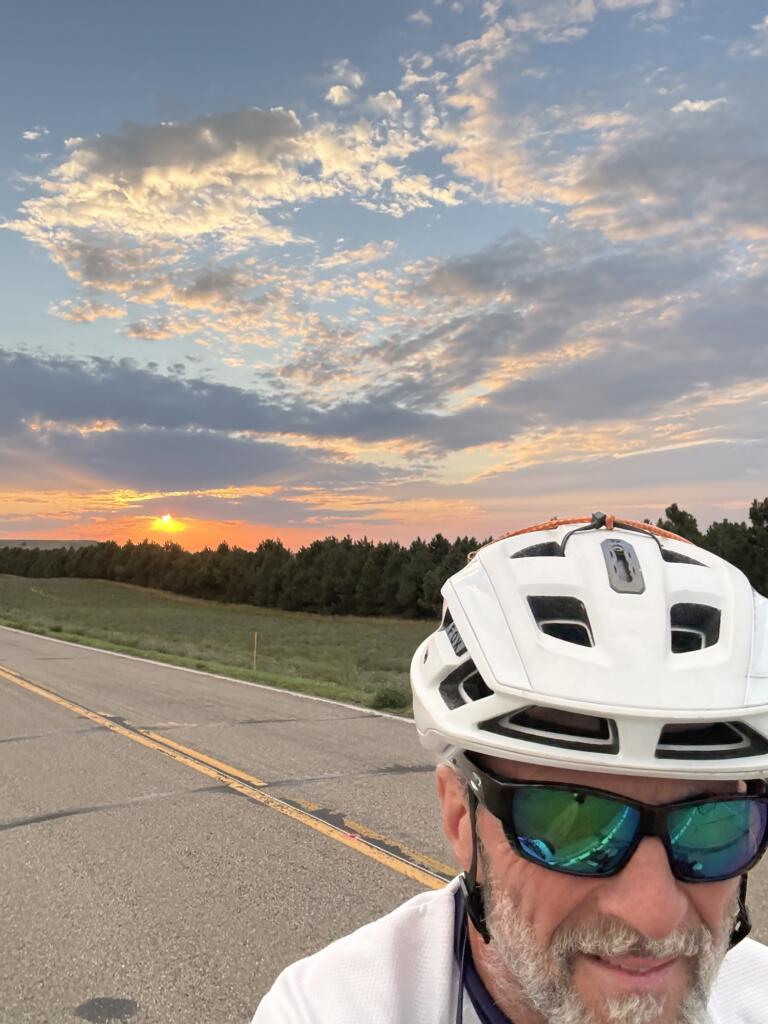 A white man wearing sunglasses and a bike helmet rides takes a selfie with a sunset behind him.