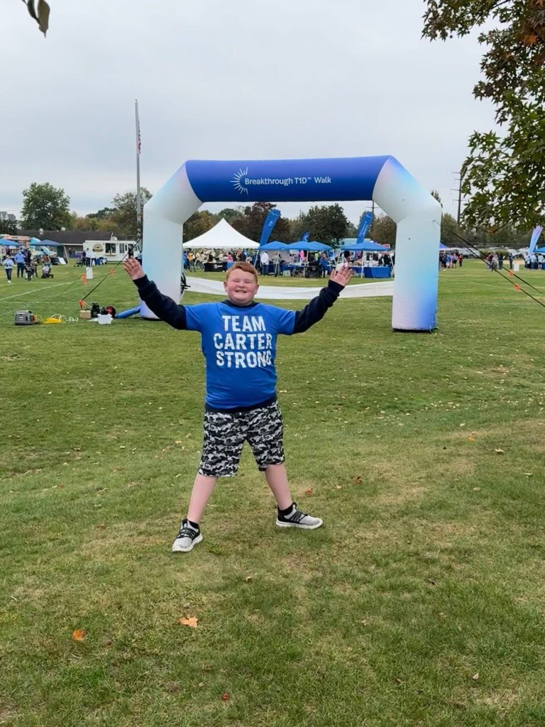 A white boy poses with his arms raised in the air while wearing a t-shirt reading "Team Carter Strong" in front of a large inflatable gate reading "Breakthrough T1D Walk."