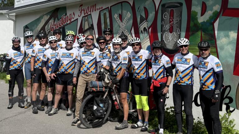 20 men in cycling gear and bike helmets, all wearing identical jerseys reading "Ride Pat Ride," pose for a photo with a bike in front of a mural reading "Greetings from Fairport, NY."