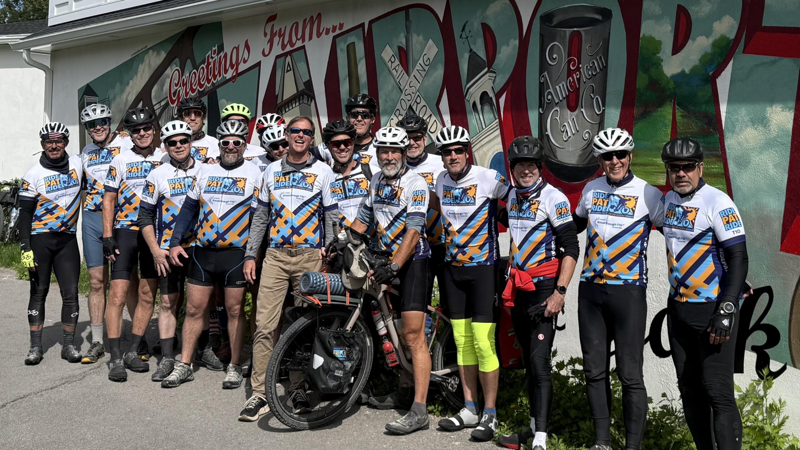 20 men in cycling gear and bike helmets, all wearing identical jerseys reading "Ride Pat Ride," pose for a photo with a bike in front of a mural reading "Greetings from Fairport, NY."
