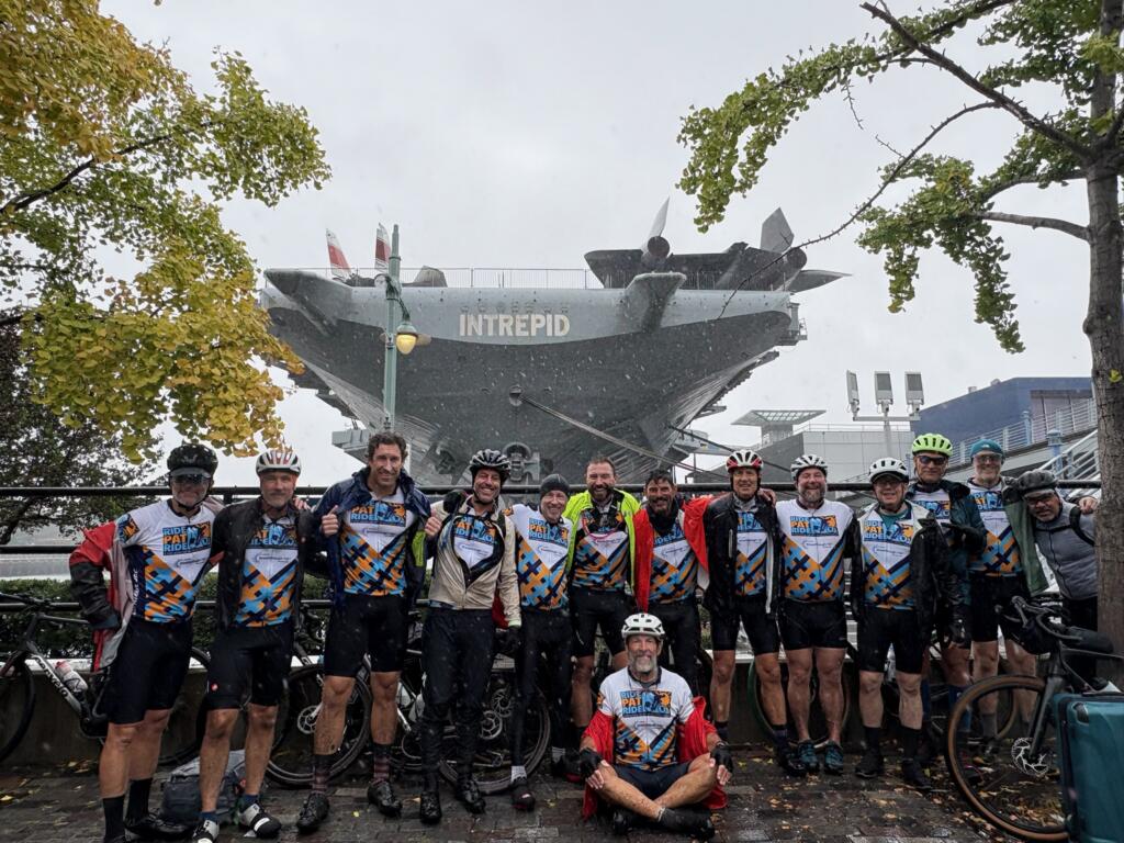 20 white men wearing matching cycling jerseys reading "Ride Pat Ride" pose for a photo in New York City in front of an aircraft carrier named "Intrepid."