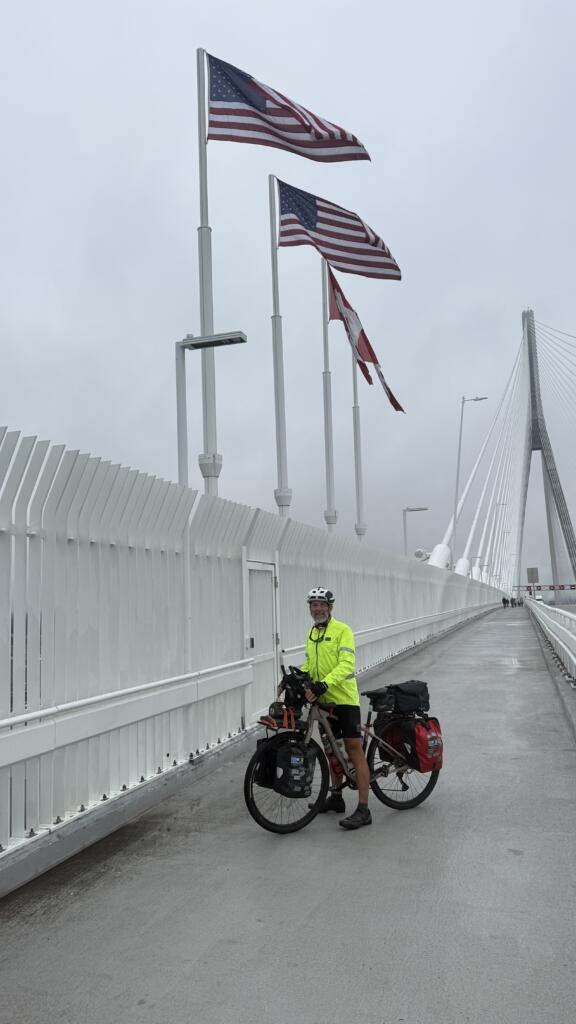 A white man in a hi-vis vest poses with his bike on a new suspension bridge beneath American and Canadian flags.
