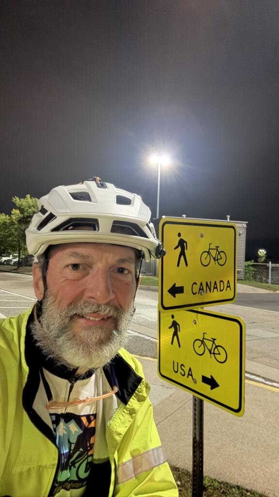 A white man wearing a bicycle helmet and hi-vis vest smiles next to signs pointing in different directions for bicycle and pedestrian traffic to "Canada" and "USA."