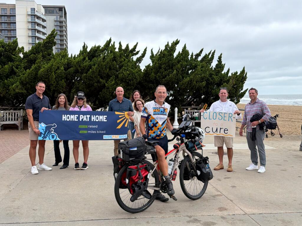 A group of people standing on a beach, including a man straddling a bicycle laden with saddlebags, stand while holding signs reading "Home Pat Home" and "Closer for a Cure."