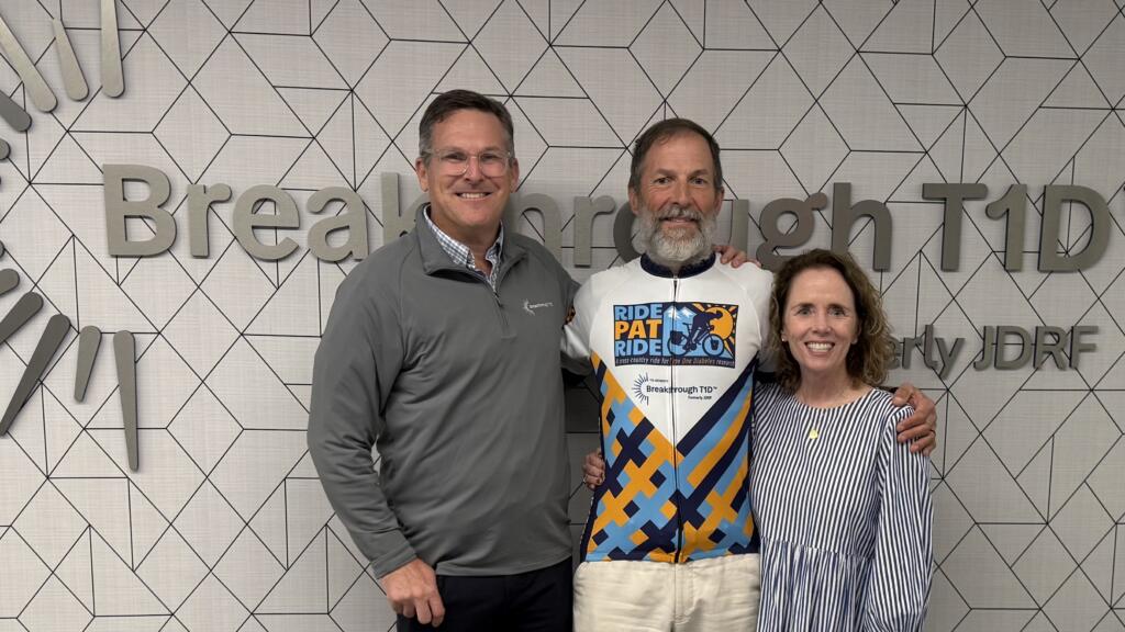 Two white men and a white woman smile while standing arm-in-arm in front of a large sign reading "Breakthrough T1D."